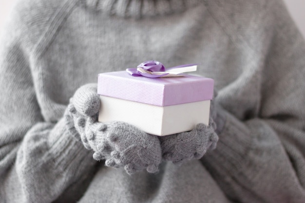 Two hands in gray knitted mittens hold a small gift box against the background of a gray knitted sweater