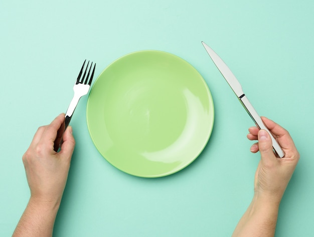 Two hands hold a metal knife and fork on the surface of a round empty green plate, top view