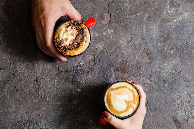 Two hands holding cups of hot coffee on dark background, top view