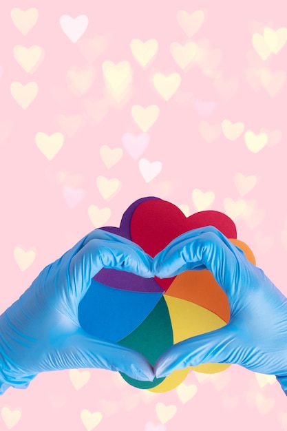 Two hands in protective blue gloves show a heart shape over an iridescent paper circle on a pink background