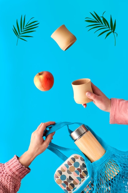 Two hands with cup and palm leaf, net bag with lunch box and tea flask flying. Zero waste tea in in eco friendly insulated bamboo flask. Zero waste concept, surreal levitation on blue wall