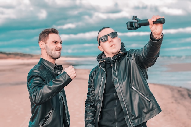 Two Handsome Male Friends Making Selfie Using Action Camera with Gimbal Stabilizer at the Beach.