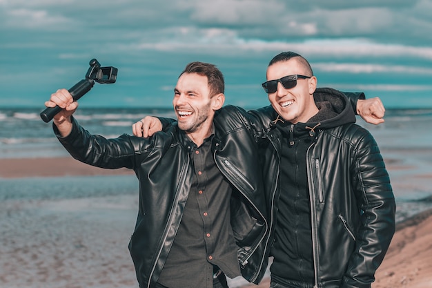 Due amici sorridenti belli che fanno selfie utilizzando la macchina fotografica di azione con stabilizzatore cardanico in spiaggia