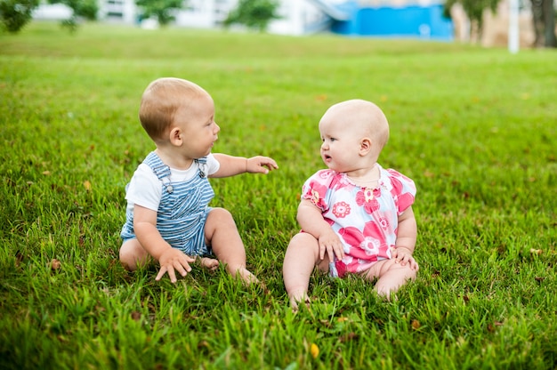 Two happy baby boy and a girl age 9 months old, sitting on the grass and interact, talk, look at each other.