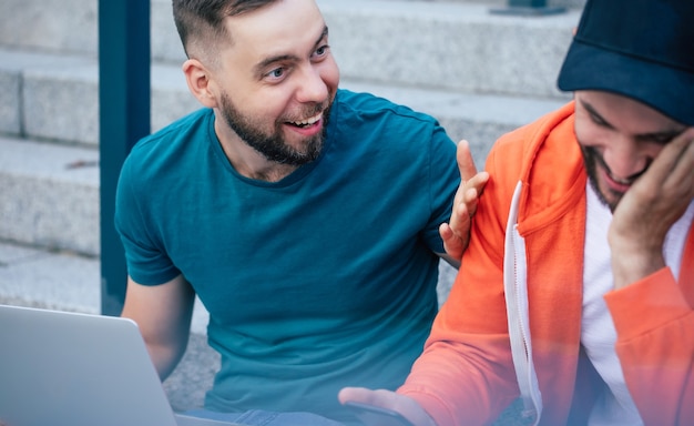 Two happy bearded hipster friends with a laptop in casual clothes are sitting on the stairs outdoor and have a conversation.