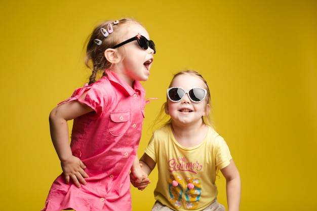 Two happy beautiful stylish little girl wearing sunglasses posing isolated yellow studio background