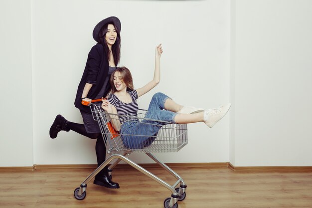 Two happy beautiful teen women  driving shopping cart indoors