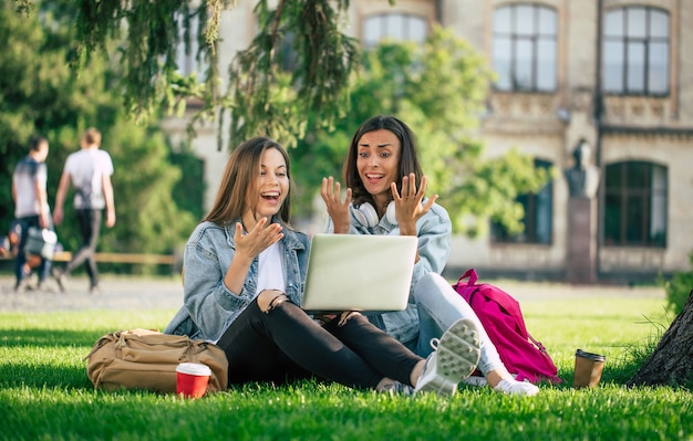 Two happy beautiful young student girl friends in casual denim clothes are relaxing in college park with laptop and smartphone by university and drinking coffee.