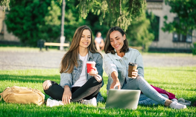 Two happy beautiful young student girl friends in casual denim clothes are relaxing in college park with laptop and smartphone by university and drinking coffee.