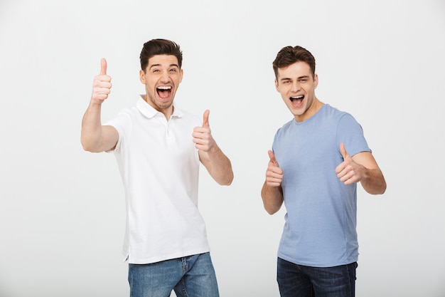 Two happy buddies 30s wearing casual t-shirt and jeans smiling and showing thumbs up on camera, isolated over white background