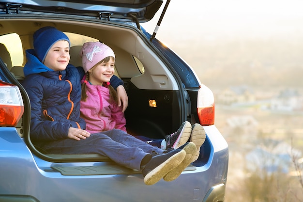 Two happy children boy and girl sitting together in a car trunk. Cheerful brother and sister hugging each other in family vehicle luggage compartment.