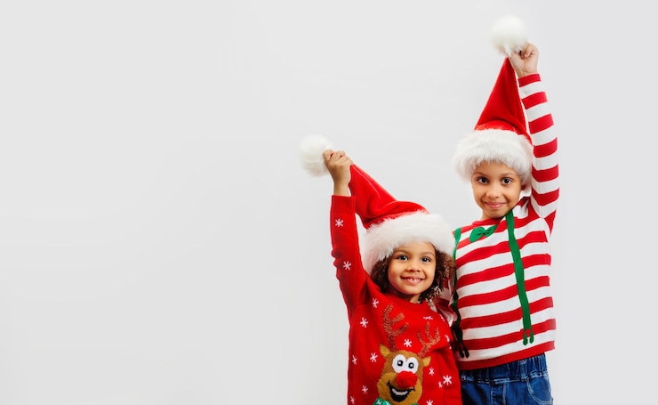Photo two happy children in christmas costumes hold up red santa hats in greeting on white background