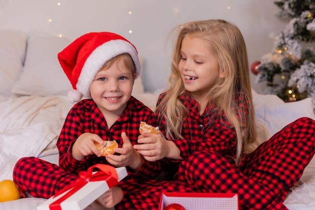 Due bambini felici in pigiama per natale mangiano mandarini e giocano con i giocattoli di natale