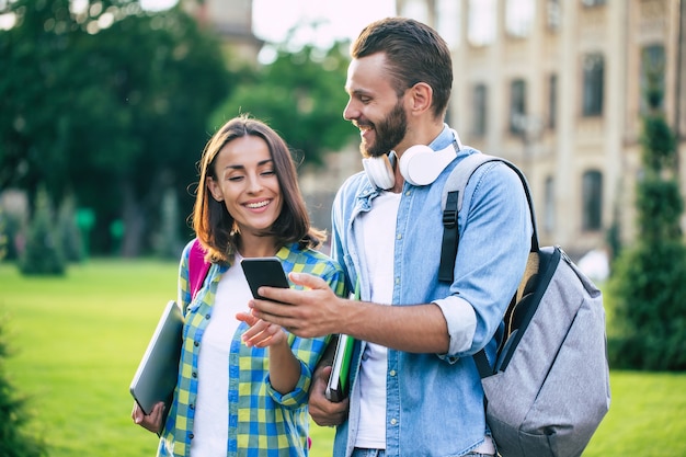 배낭과 스마트 폰을 든 두 명의 행복하고 흥분된 학생 친구가 대화를 나누고 야외에서 걷고 있습니다. 대학에서 수업 후 아름다운 커플