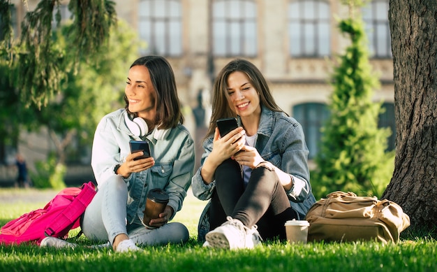 カジュアルなデニムの服を着た2人の幸せで興奮した学生のガールフレンドは、スマートフォンを持って大学の公園でリラックスして楽しんでいます。