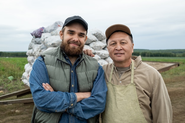 Два счастливых фермера в спецодежде стоят против кучи мешков с картофелем