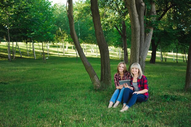 Two happy friends searching media content online sitting on the grass in a park.