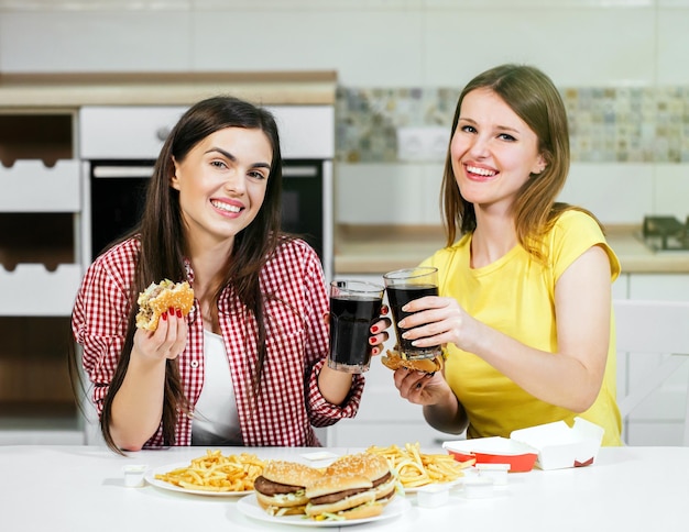 Two happy friends spending time together eating tasty fast food in home kitchen having nice pleasant talk