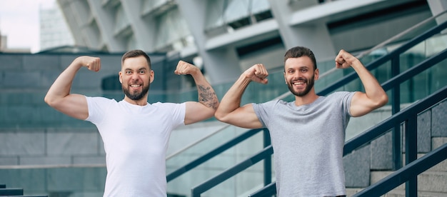 Two happy funny excited handsome bearded best friends in casual t-shirts are showing biceps on camera and having fun. Sport, fitness, strong men, power in the team.