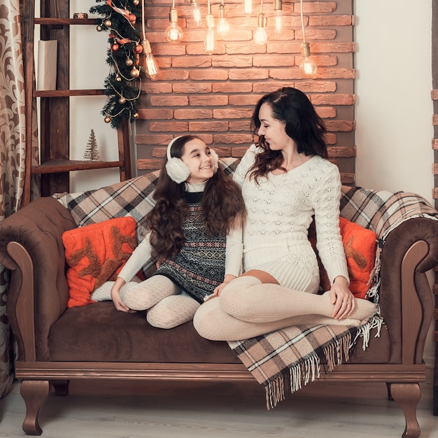 Two happy girls, mother and daughter siting on sofa in Christmas decorated room.