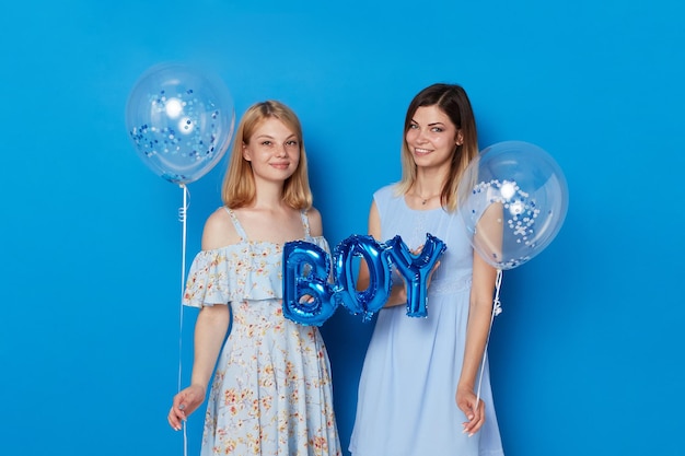 Two happy girls posing in a studio holding blue balloons and balloon with the inscription boy blue background