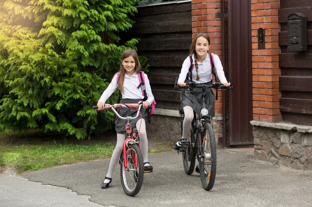 Two happy girls in school uniform with bags riding to school on bicycles