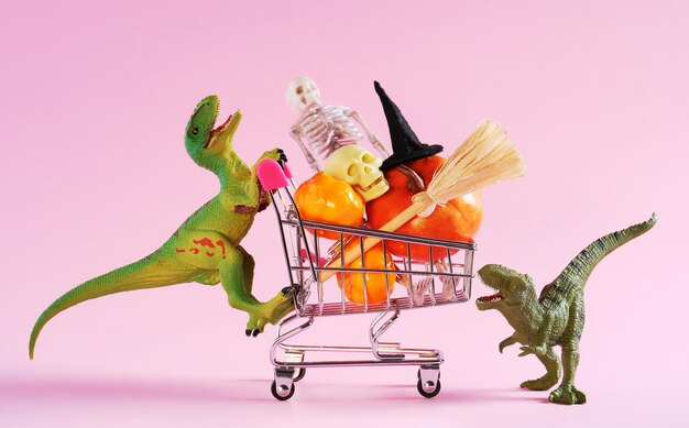 Two happy green dinosaur with shopping trolley full of halloween decorations on pink background