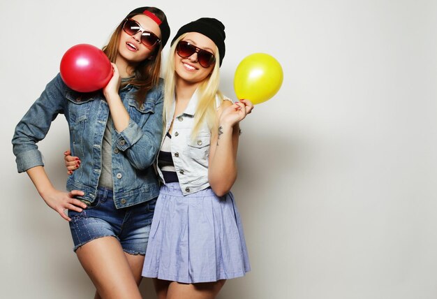 Two happy hipster girls smiling and holding colored balloons over white background