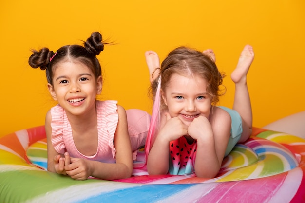 Two happy little girl in swimming suit lying on colorful inflatable mattress lollipop on yellow surface.