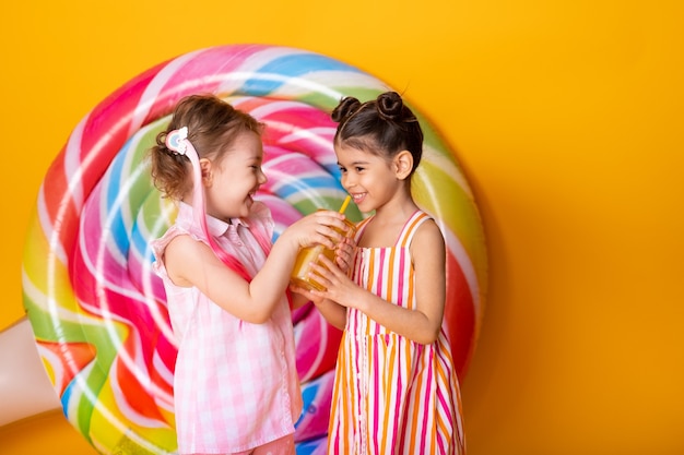 Two happy little girls in colorful dress drinking orange juice having fun on yellow surface with lollipop.