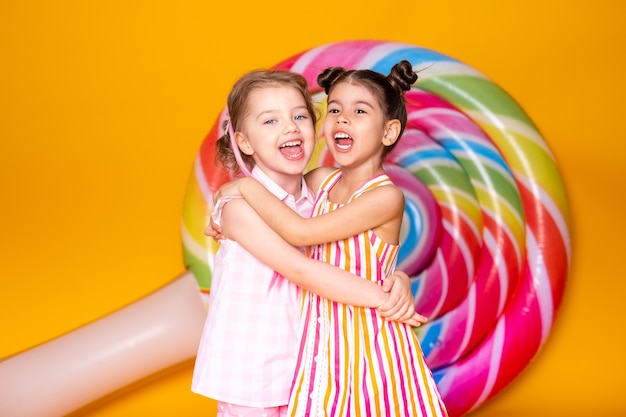 Two happy little girls in colorful dress laughing hugging having fun on yellow surface with lollipop