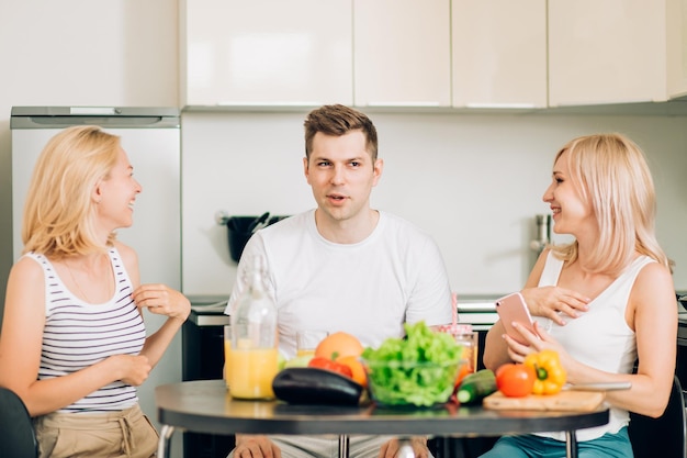 Two happy lovely women and handsome man sitting in kitchen, smiling looking at camera. Friends going to cook dinner from vegetables. Lifestyle, dieting and healthy food concept.