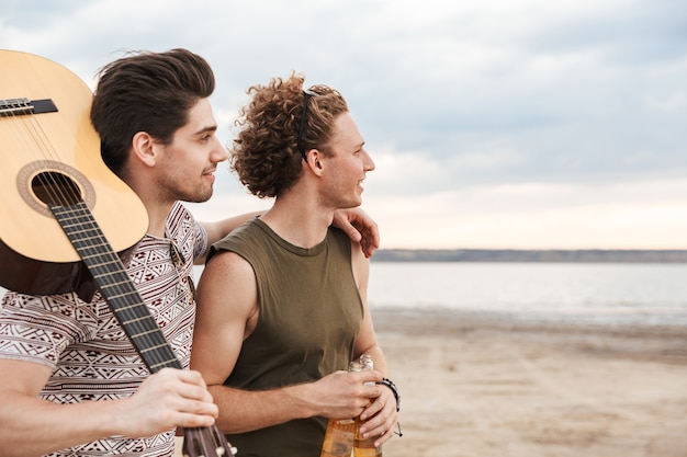 Two happy male friends walking at the beach, carrying guitar