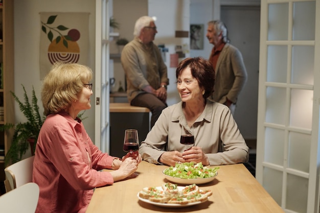 Two happy senior women with glasses of red wine having talk by served table