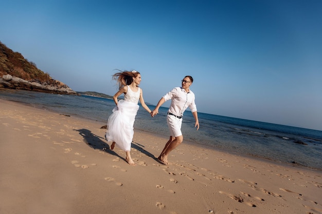 Two happy and shining lovers in white clothes, holding hands, running along the sandy seashore and looking at each other with charm. Concert of love