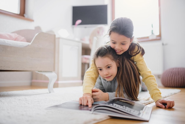 Two happy small girls sisters indoors in bedroom at home, reading a book.