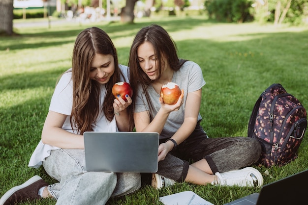 두 명의 행복한 미소를 짓고 있는 여학생들이 공원에 책과 노트북을 들고 공원에 앉아 사과를 먹고 공부하고 시험을 준비하고 있습니다. 원격 교육. 부드러운 선택적 초점입니다.