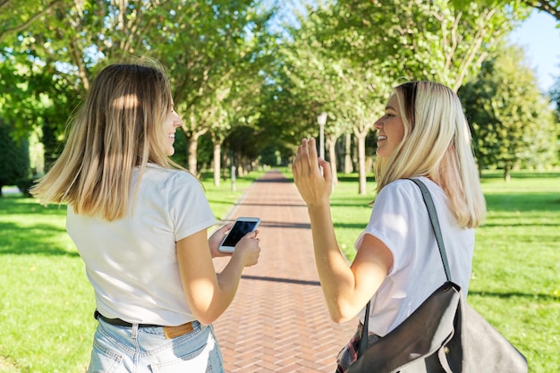 Two happy smiling talking girls teenagers students walking together, back view, young women with backpack, sunny day in the park background