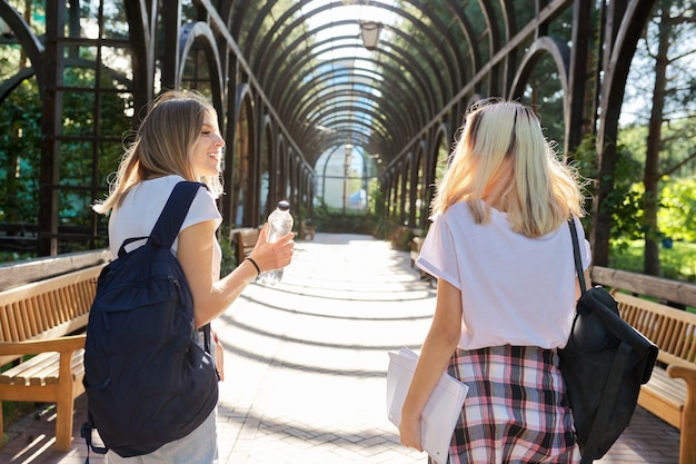 Two happy smiling talking girls teenagers students walking together, young women with backpacks, sunny day in the park background, back view