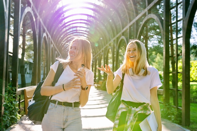 Two happy smiling talking girls teenagers students walking together, young women with backpacks, sunny day in the park background