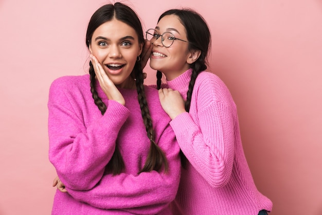 two happy teenage girls with braids in casual clothes gossiping together isolated over pink wall