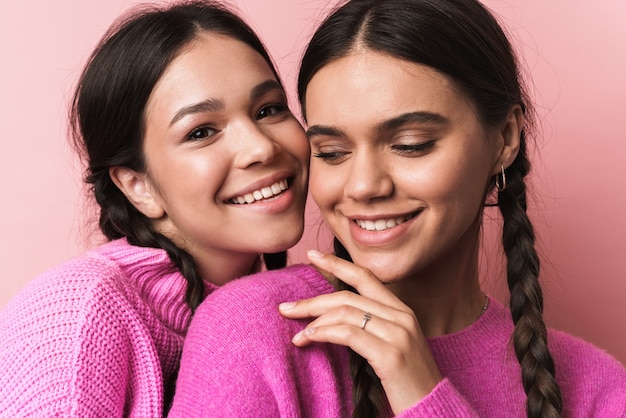 two happy teenage girls with braids in casual clothes smiling at camera isolated over pink wall