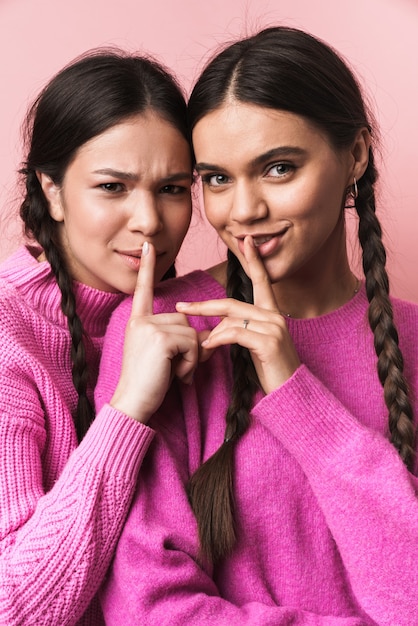 two happy teenage girls with braids smiling and keeping fingers at lips isolated over pink wall