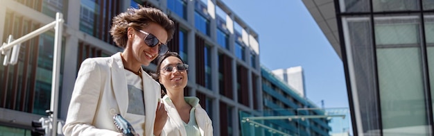 Two happy woman in trendy outfits laugh and walk outside on background of modern building