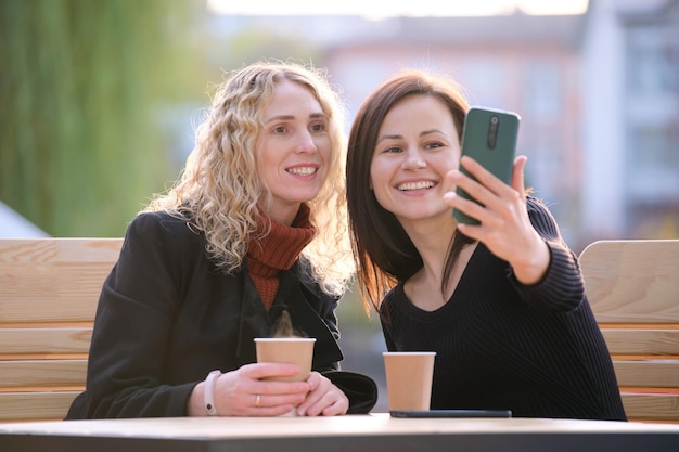 Two happy women making selfie with mobile phone sitting at city\
street cafe female friends enjoying time together outdoors