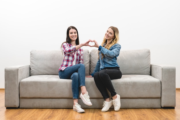 Две счастливые женщины показывают символ сердца на белом фоне стены