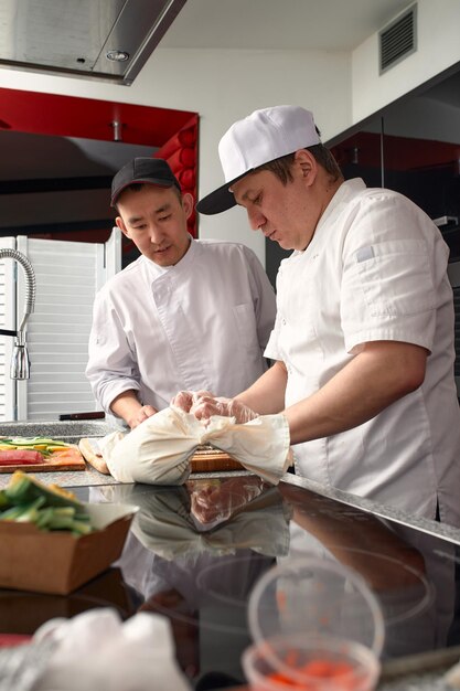 Two happy young asian chefs dressed in white uniform preparing traditional japanese sushi set in interior of modern professional kitchen