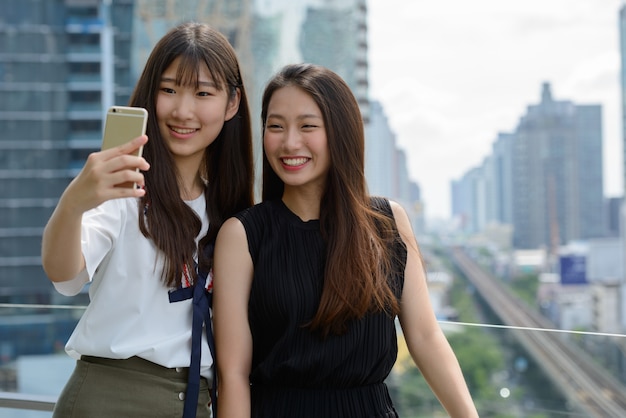 Two happy young beautiful Asian teenage girls taking selfie together against view of the city