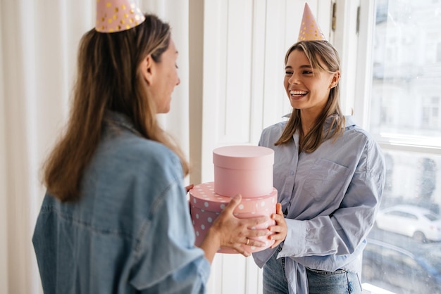 Two happy young caucasian female friends in party caps exchange gifts while standing at window. Birthday concept.