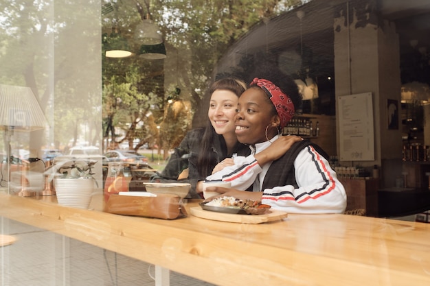 Две счастливые молодые подруги в повседневной одежде сидят за обслуживаемым столиком в кафе